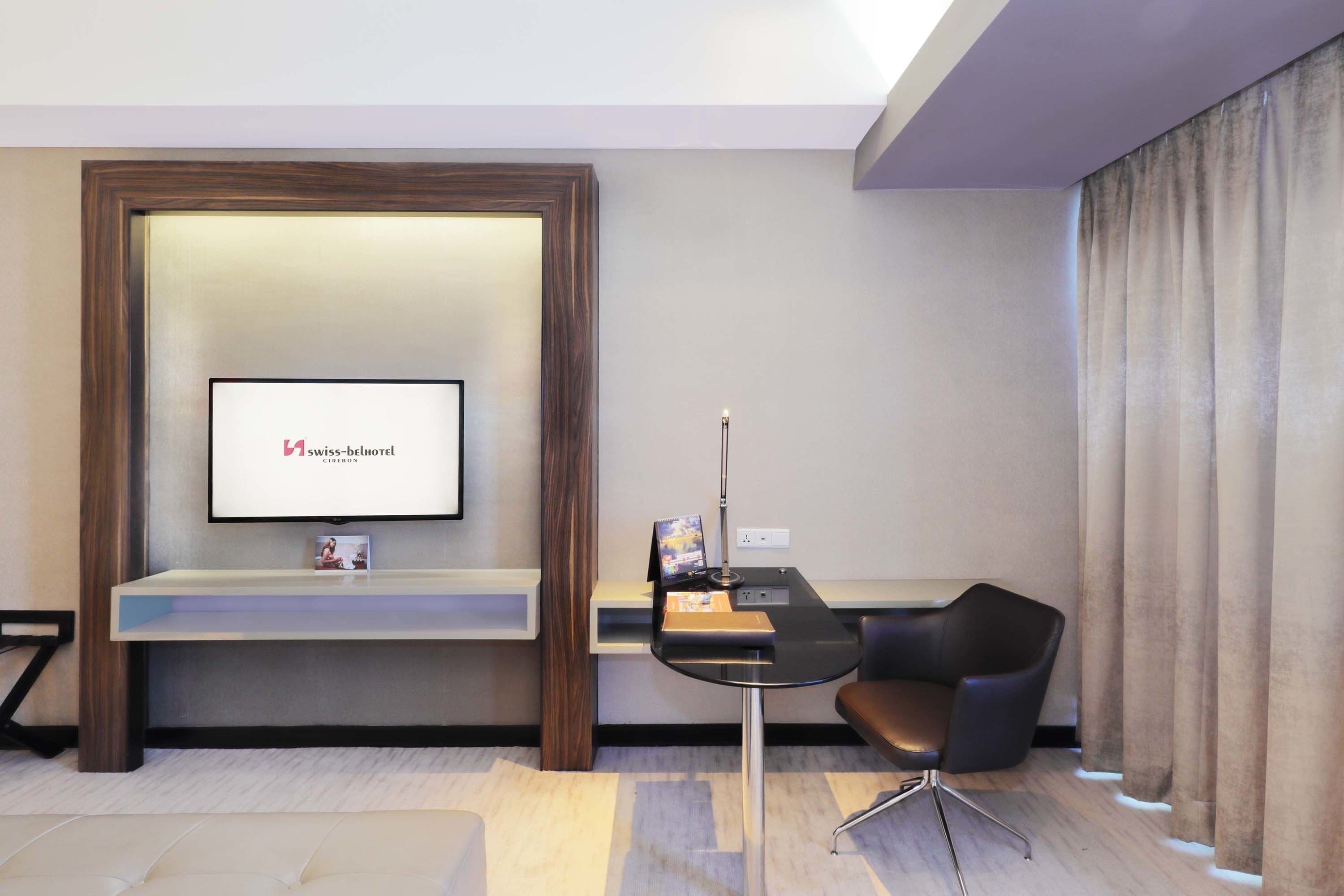 Un espace d’accueil moderne, avec un téléviseur à écran plat affichant le logo de Swiss-Belhotel, un comptoir d’accueil, un fauteuil noir et une petite table avec une lampe.