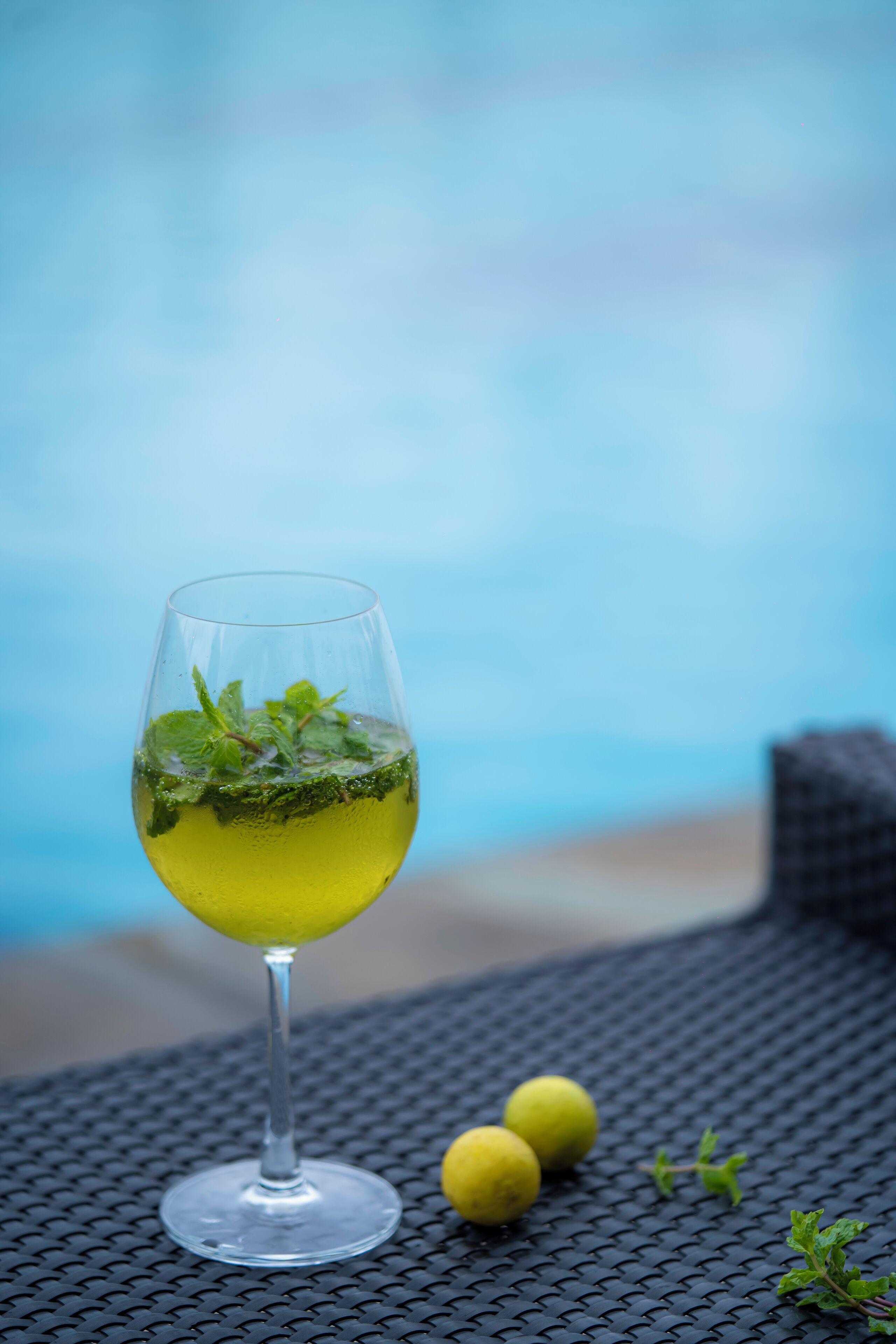 A glass of yellow beverage with mint leaves, two lemons, and a small sprig of mint on a textured surface near a pool.