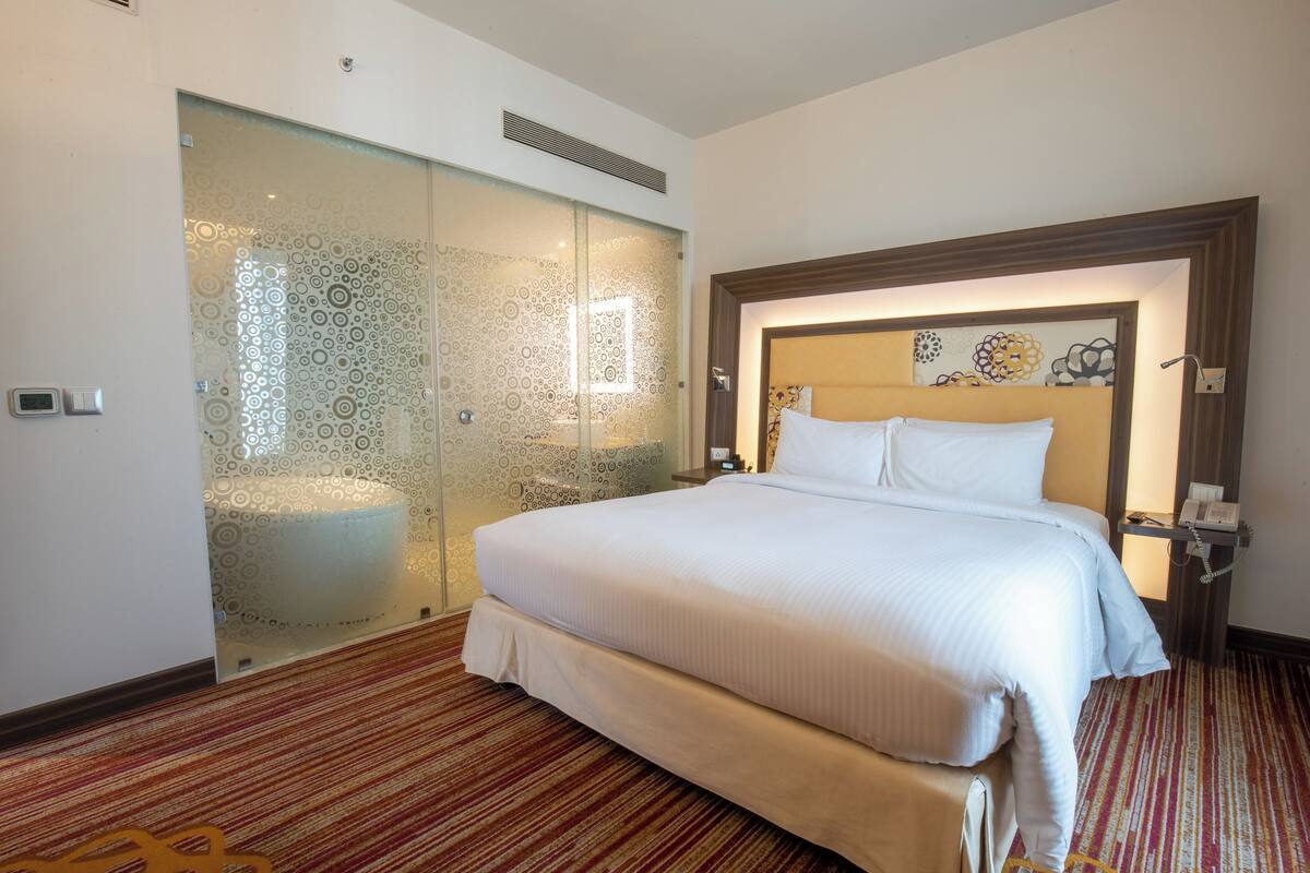 A hotel room with a large bed, a glass partition wall, and a patterned carpet.