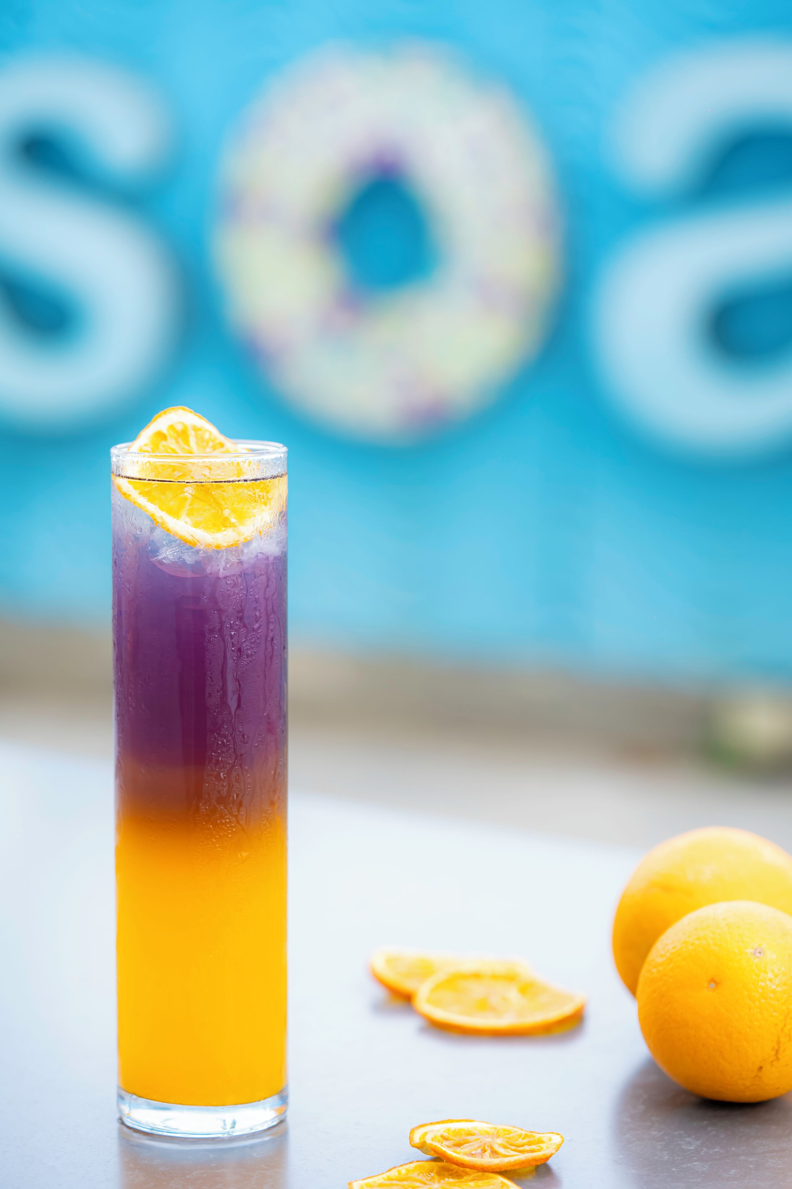 A tall glass with a layered drink, a slice of orange on top, and two whole oranges on the side.