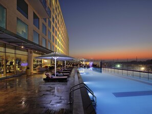 A rooftop pool with lounge chairs and umbrellas, a glass facade building, and a clear sky. - Novotel Pune Viman Nagar Hotel (Pune)