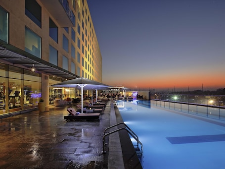 A rooftop pool with lounge chairs and umbrellas, a glass facade building, and a clear sky.