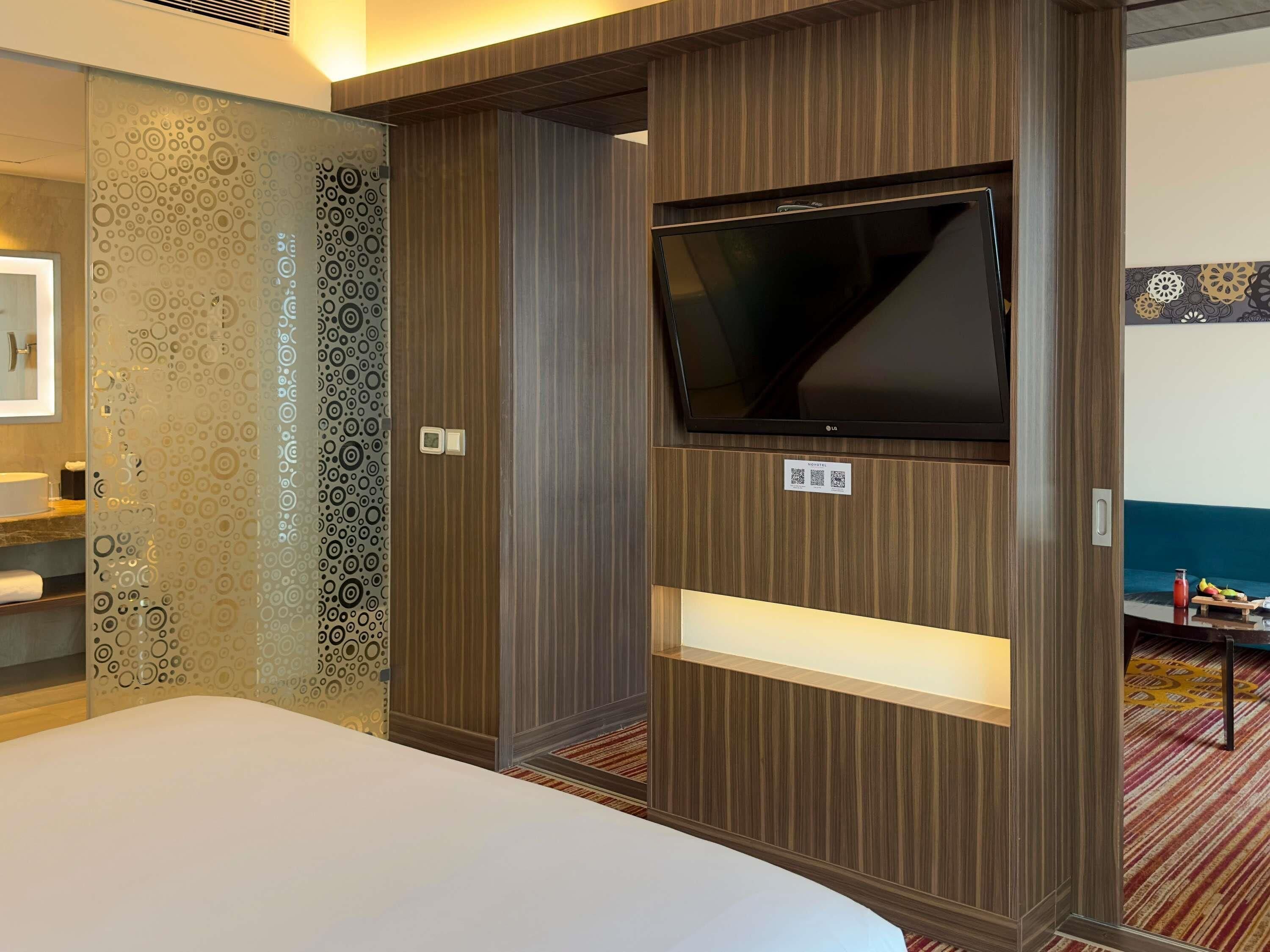 A hotel room with a bed, a TV mounted on a wooden panel, a small shelf, and a patterned wall.