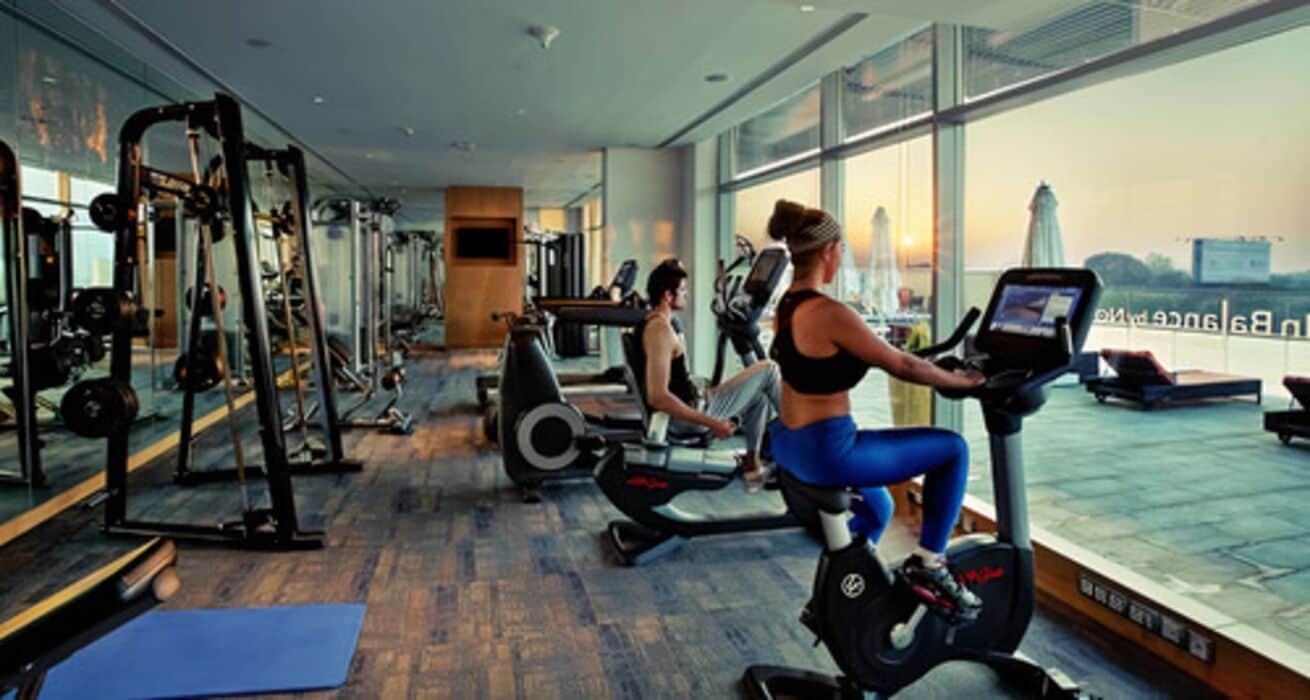 Ein Fitnessstudio mit Laufbändern, Krafttrainingsgeräten und großen Fenstern mit Blick auf die Stadt.