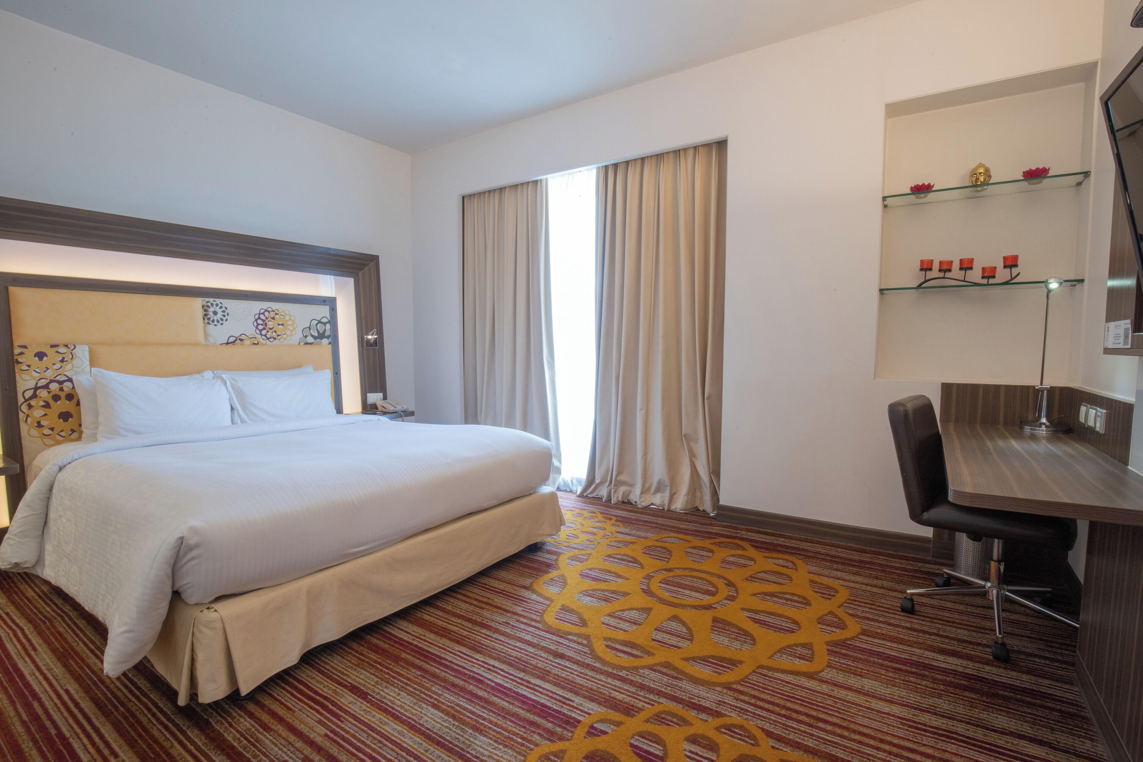 Ein Hotelzimmer mit einem großen Bett, einem Schreibtisch mit Stuhl, einem fenster mit Vorhängen und einem dekorativen Teppich.