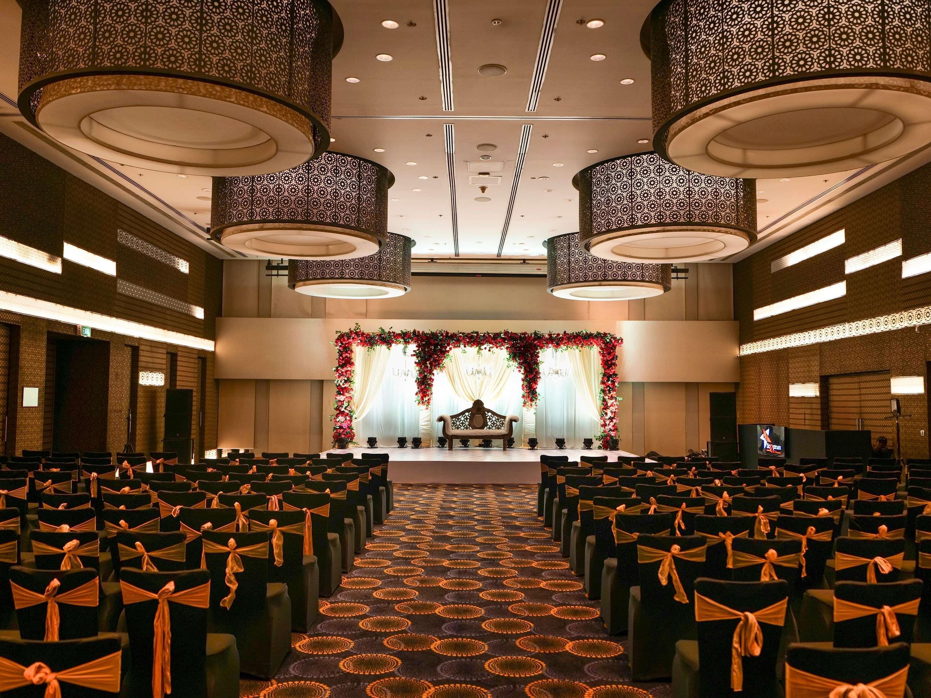 Un salón de conferencias con filas de sillas, un escenario con fondo floral y lámparas ornamentales en el techo.