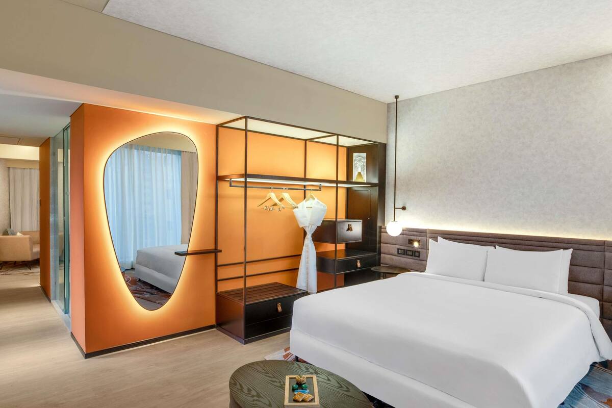 A modern hotel room with a large bed, a built-in wardrobe, a vanity with a mirror, and a bedside table with a lamp.