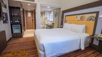 Habitación de hotel con una cama grande, teléfono en la mesita de noche, un refrigerador pequeño y un baño visible a través de una puerta de vidrio.