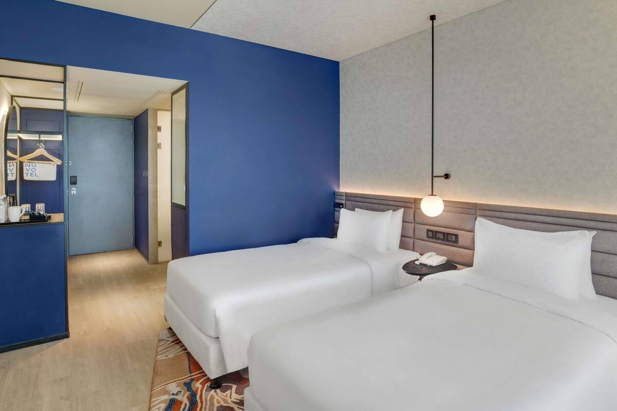 A hotel room with two beds, a bedside table, a nightstand, a hanging light fixture, and a mirror.