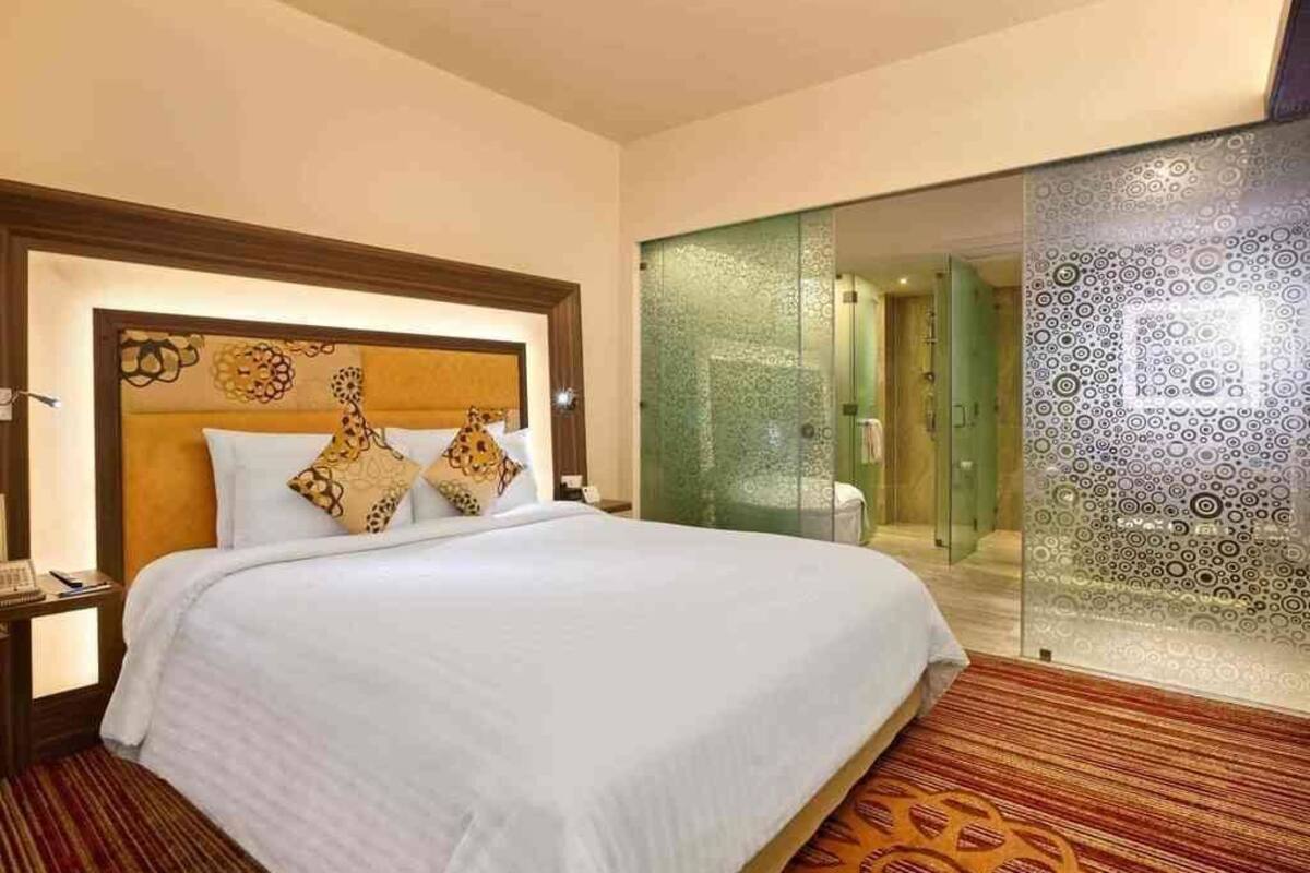 A hotel room with a large bed, a nightstand, a glass shower enclosure, and a patterned carpet.