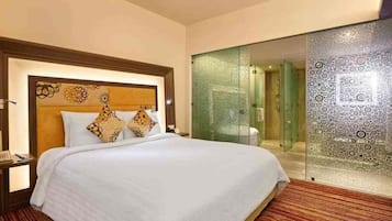 A hotel room with a large bed, a nightstand, a glass shower enclosure, and a patterned carpet.