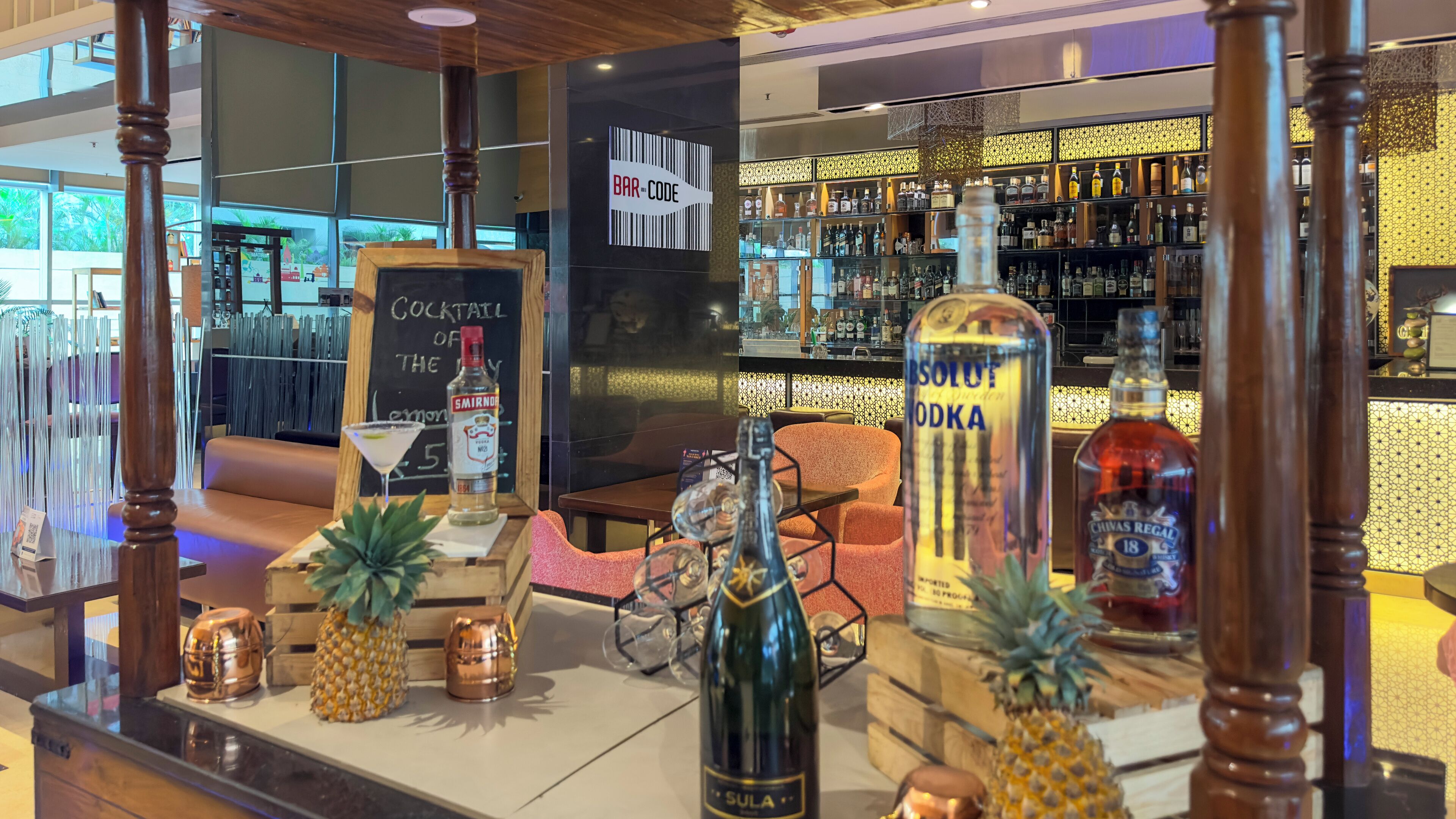 Zona de bar con mostrador de madera, varias botellas de licor, carta de cócteles y un área de estar con sofá y sillones.