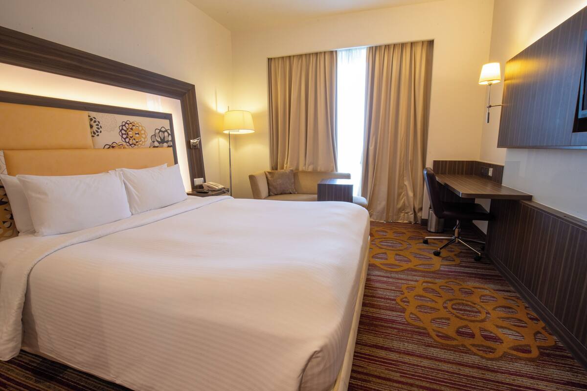 A hotel room with a large bed, a desk, a chair, a lamp, and a TV on the wall.