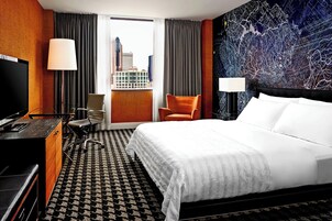 A hotel room with a large bed, a desk with a TV, a chair, and a view of the city through a window.