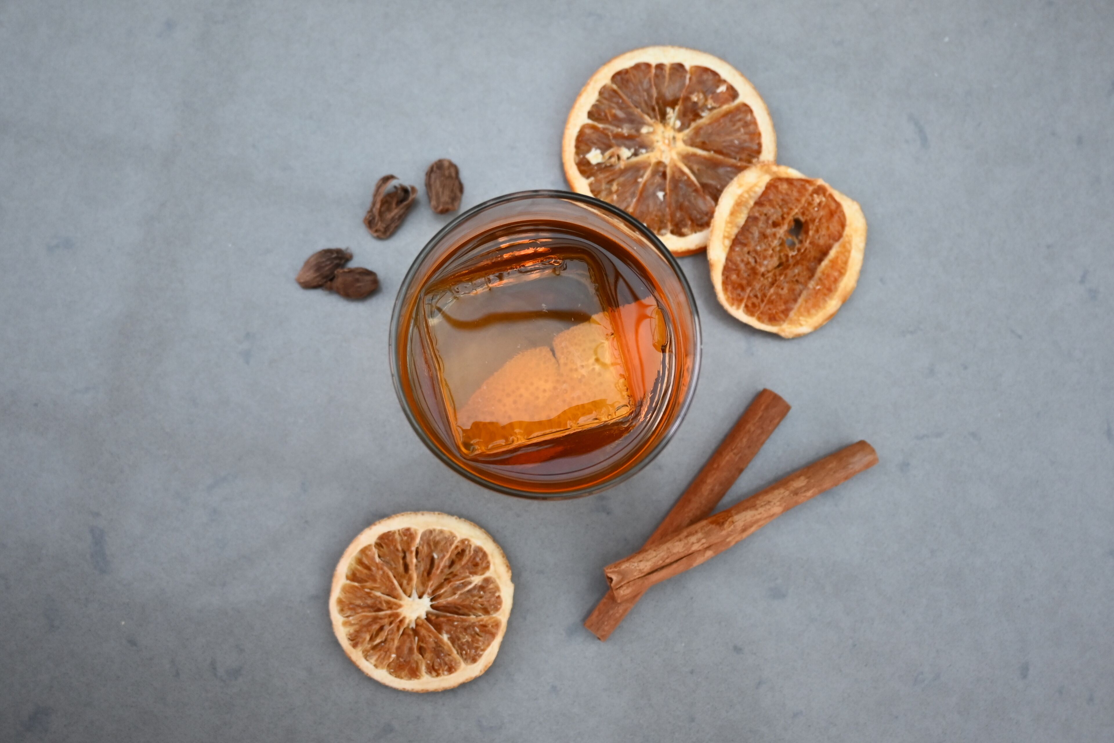 A glass of amber liquid with cinnamon sticks and dried orange slices on a grey surface.