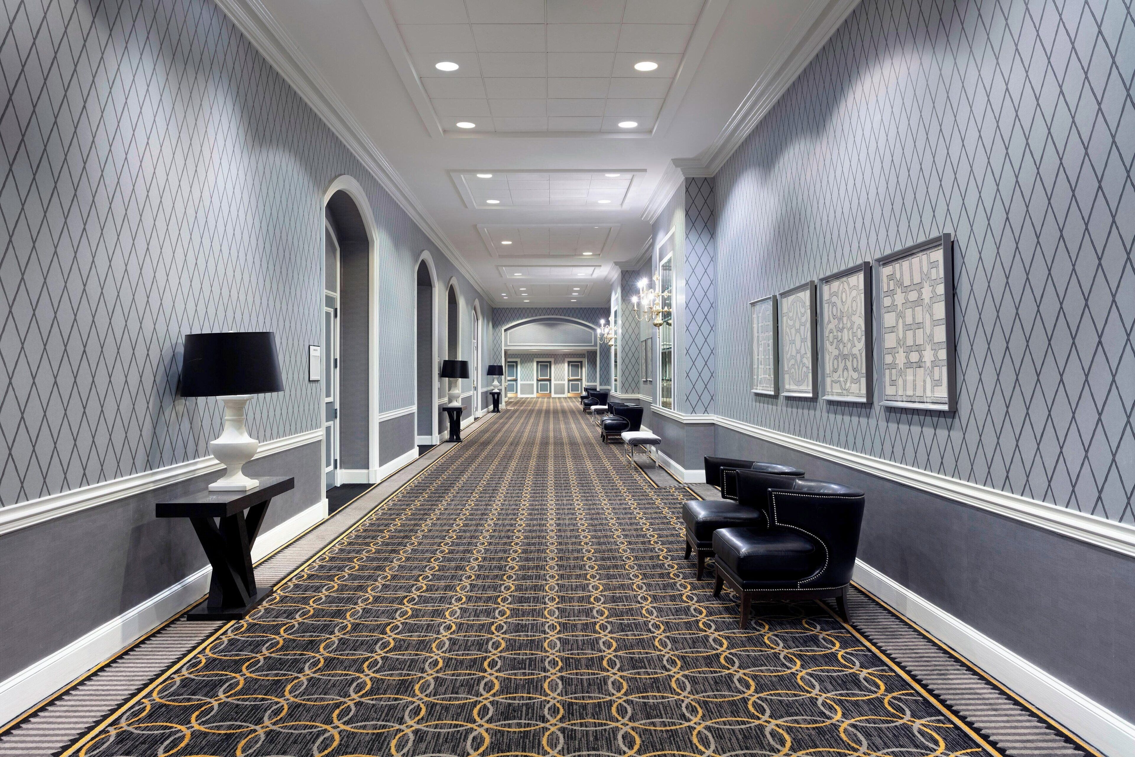 A hotel corridor with a patterned carpet, black leather seating, and framed artwork on the walls.