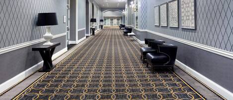 A hotel corridor with a patterned carpet, black leather seating, and framed artwork on the walls.