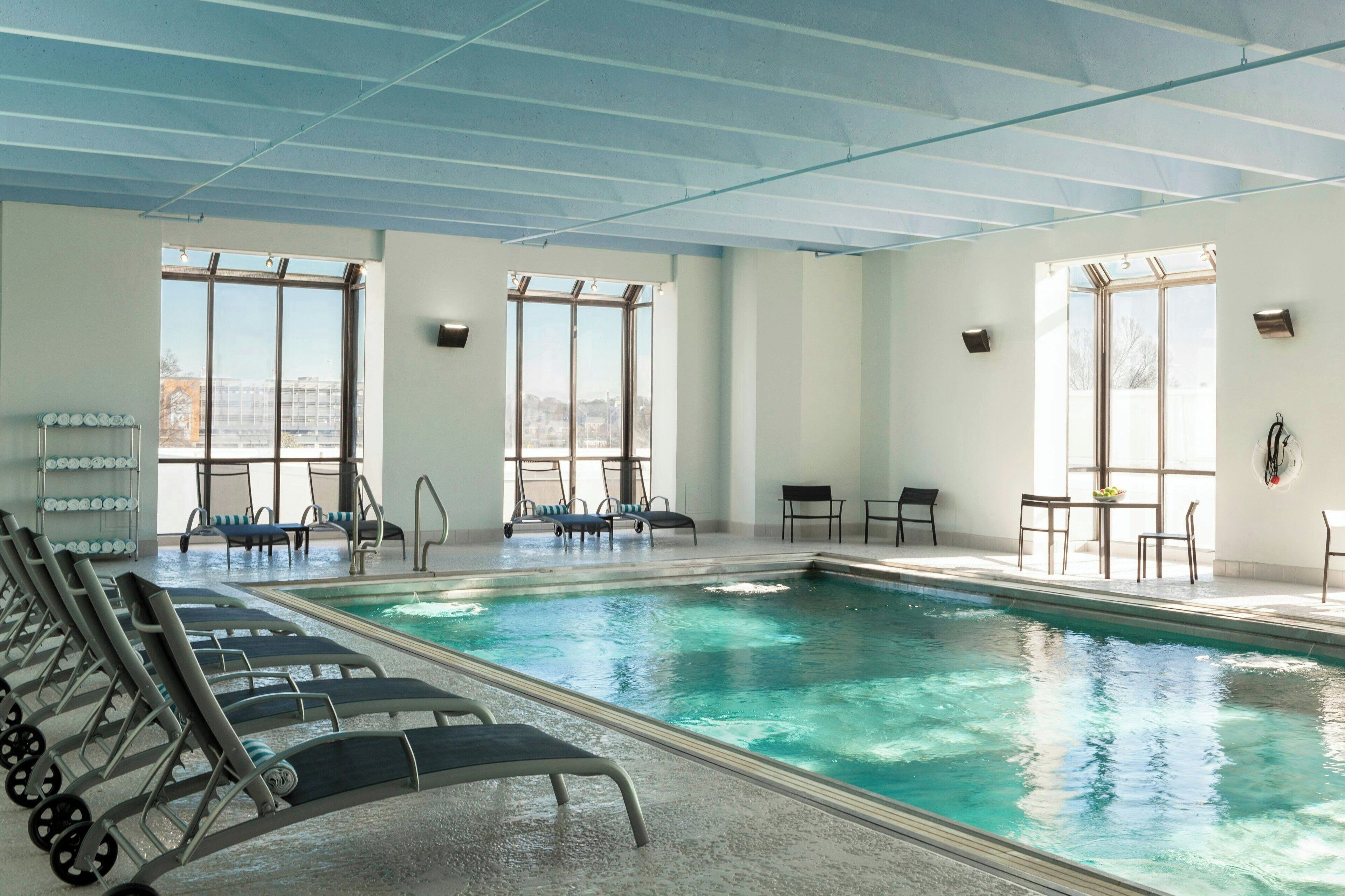 A swimming pool with lounge chairs and tables, large windows, and a view of the city.