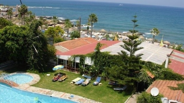 A swimming pool with a view of the ocean, surrounded by palm trees and a red-roofed building. - Anatoli Apartments (Hersonissos)