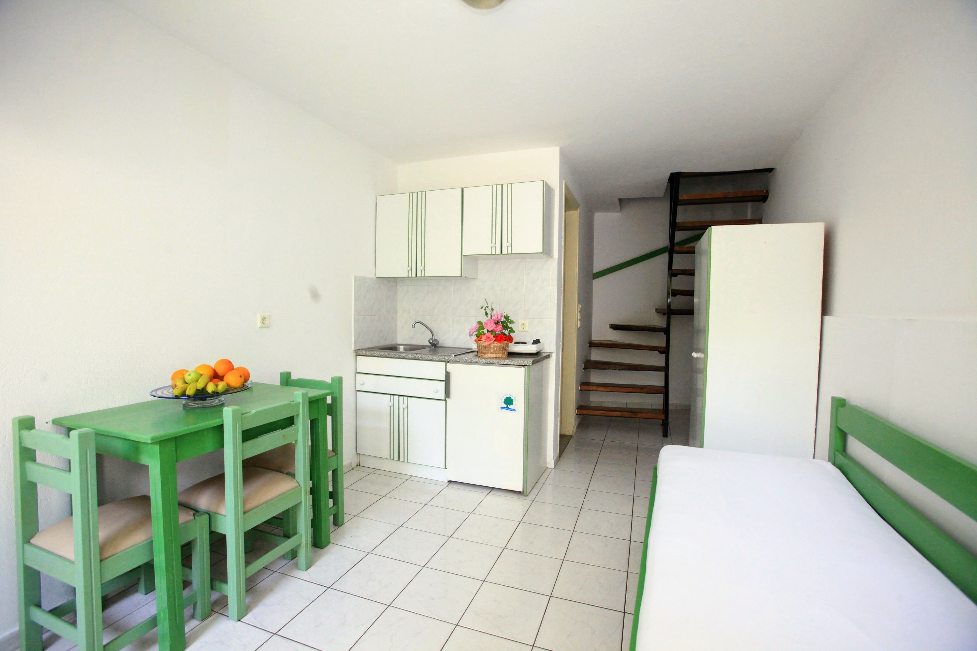 A compact room with a kitchenette, a dining table with chairs, a bed, and a staircase.