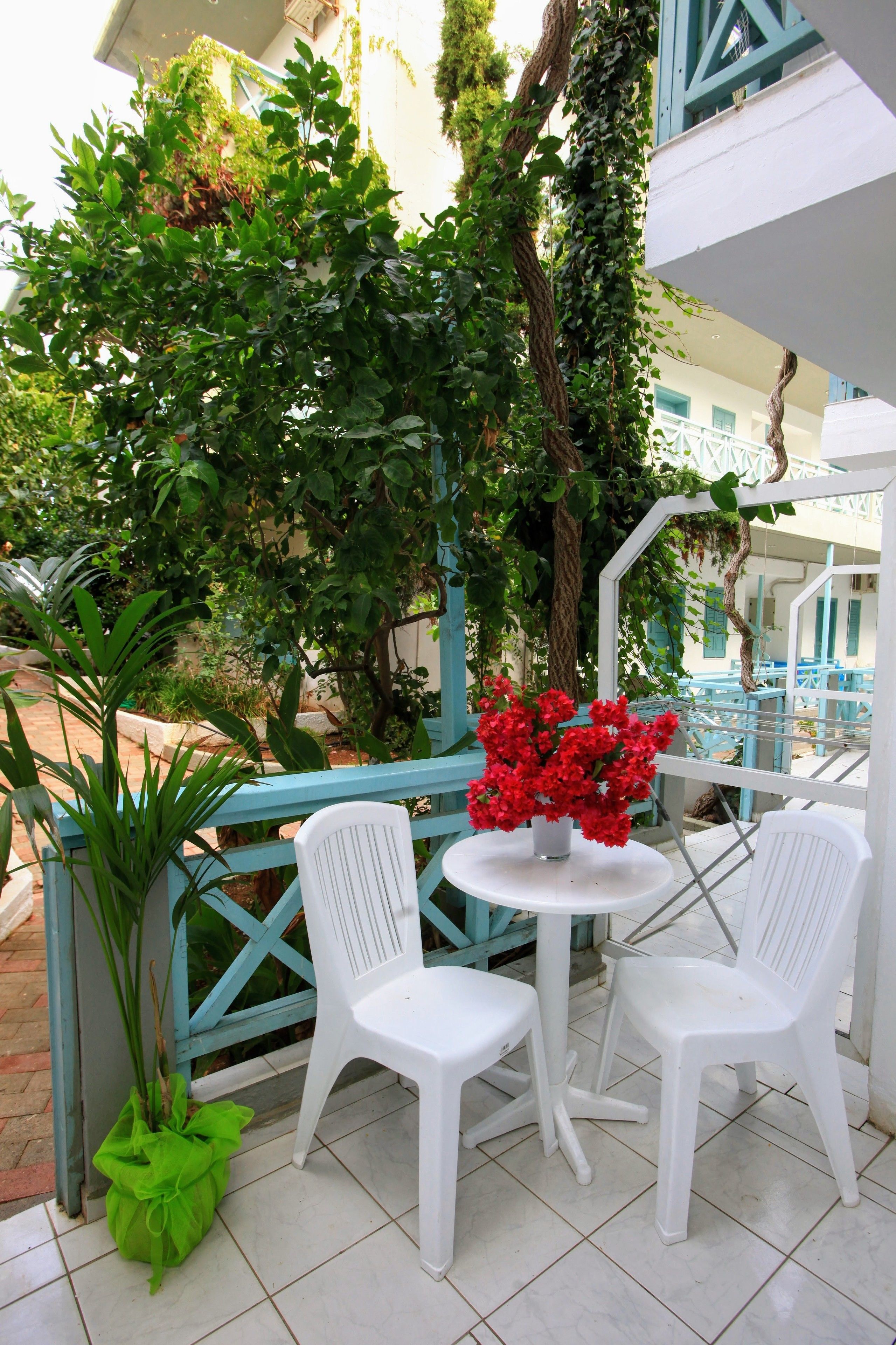 Eine Terrasse mit weißen Plastikstühlen und einem Tisch, umgeben von viel Grün und einem blauen Geländer.