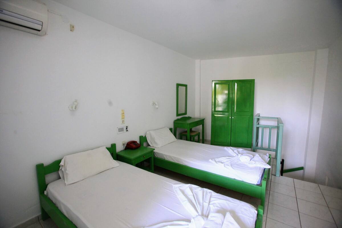 Ein Zimmer mit zwei Einzelbetten, einem grünen Kleiderschrank, einem kleinen Tisch und einem grünen Stuhl.