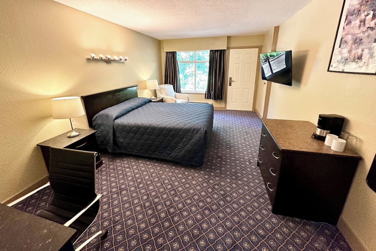 A hotel room with a bed, a desk with a coffee maker, a TV, and a window with curtains.