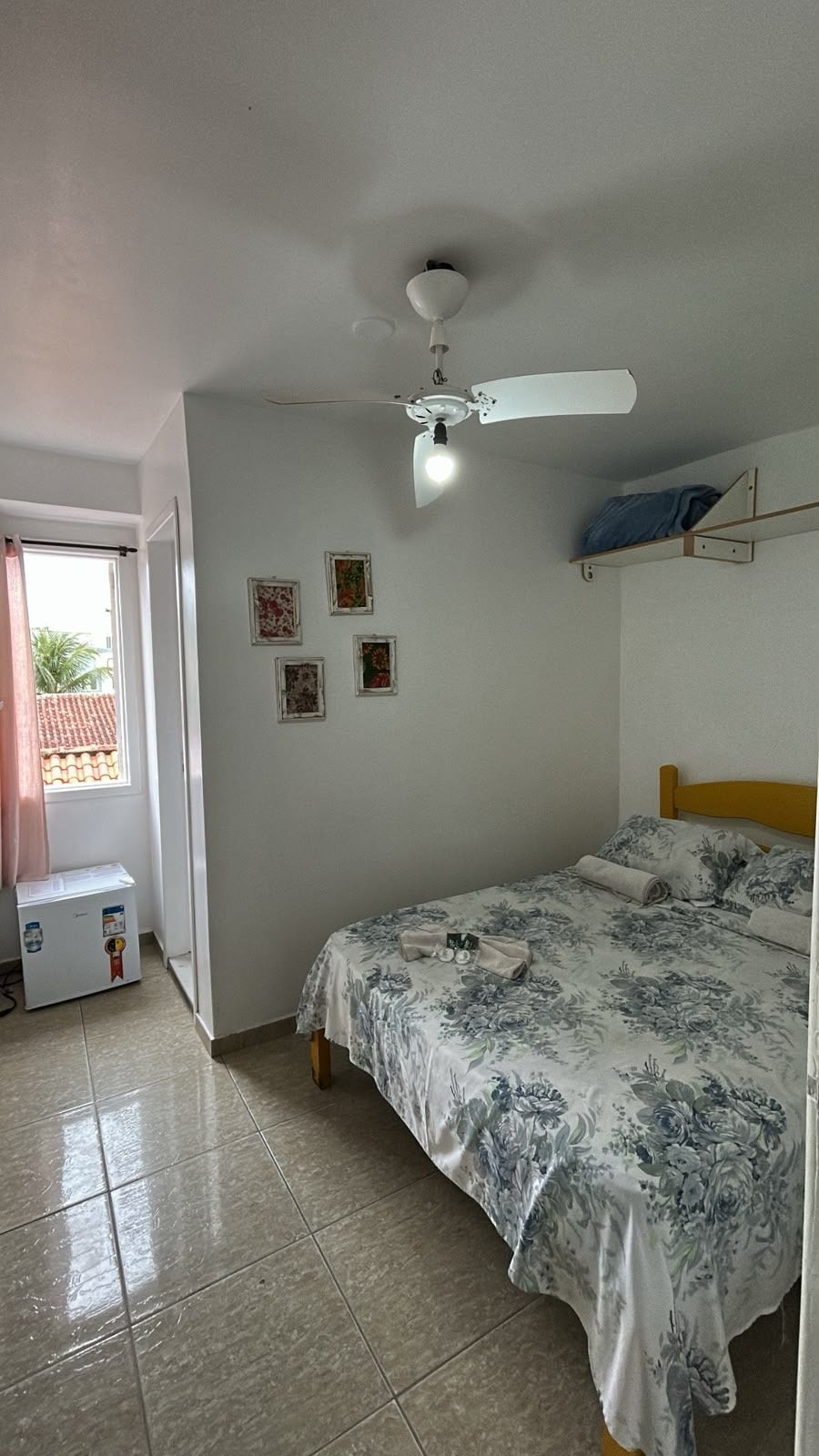 Un dormitorio con cama, ventilador de techo, ventana y estante fijado a la pared.