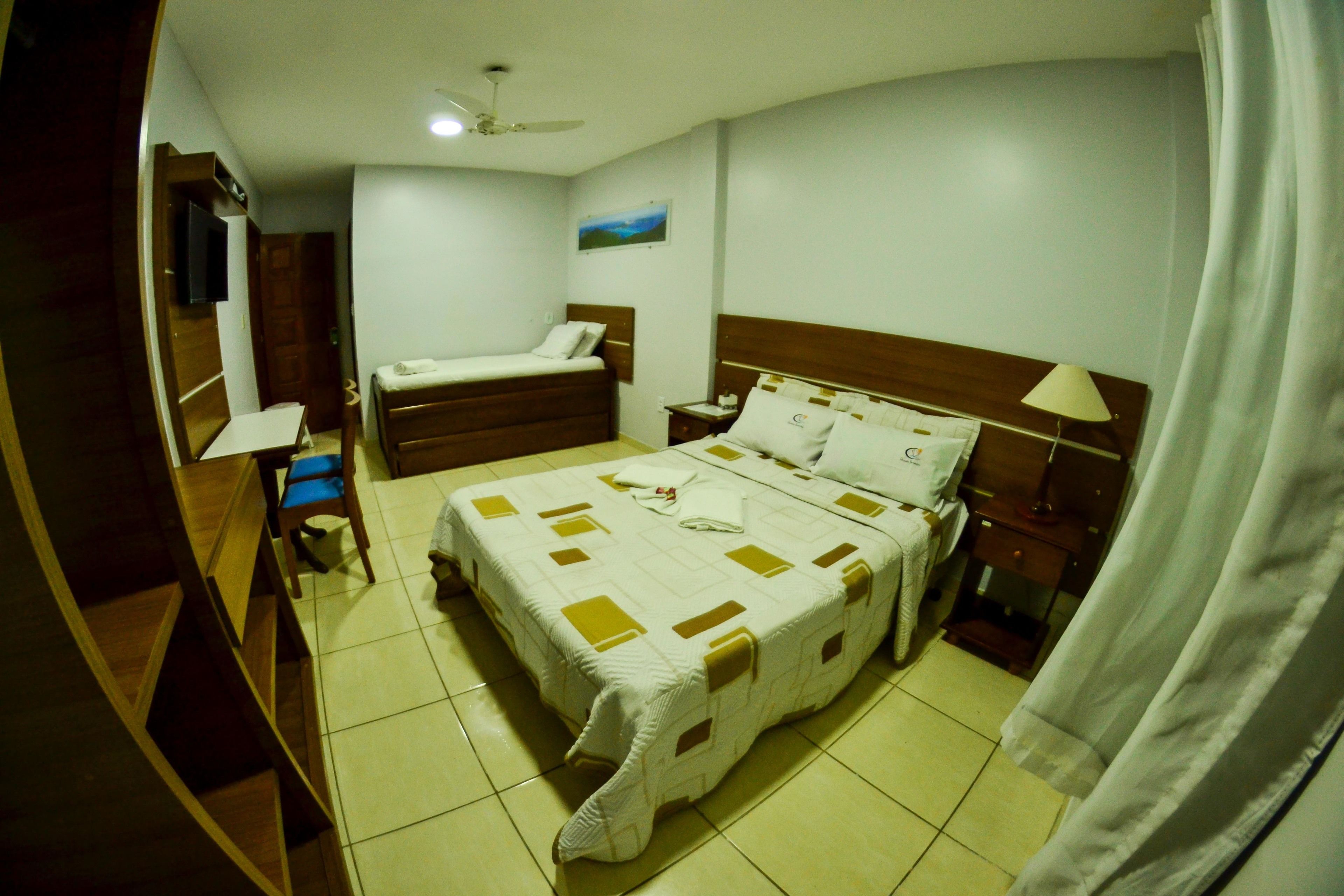 Una habitación de hotel con una cama grande, una cama más pequeña, un escritorio, una silla, un televisor y un ventilador de techo.