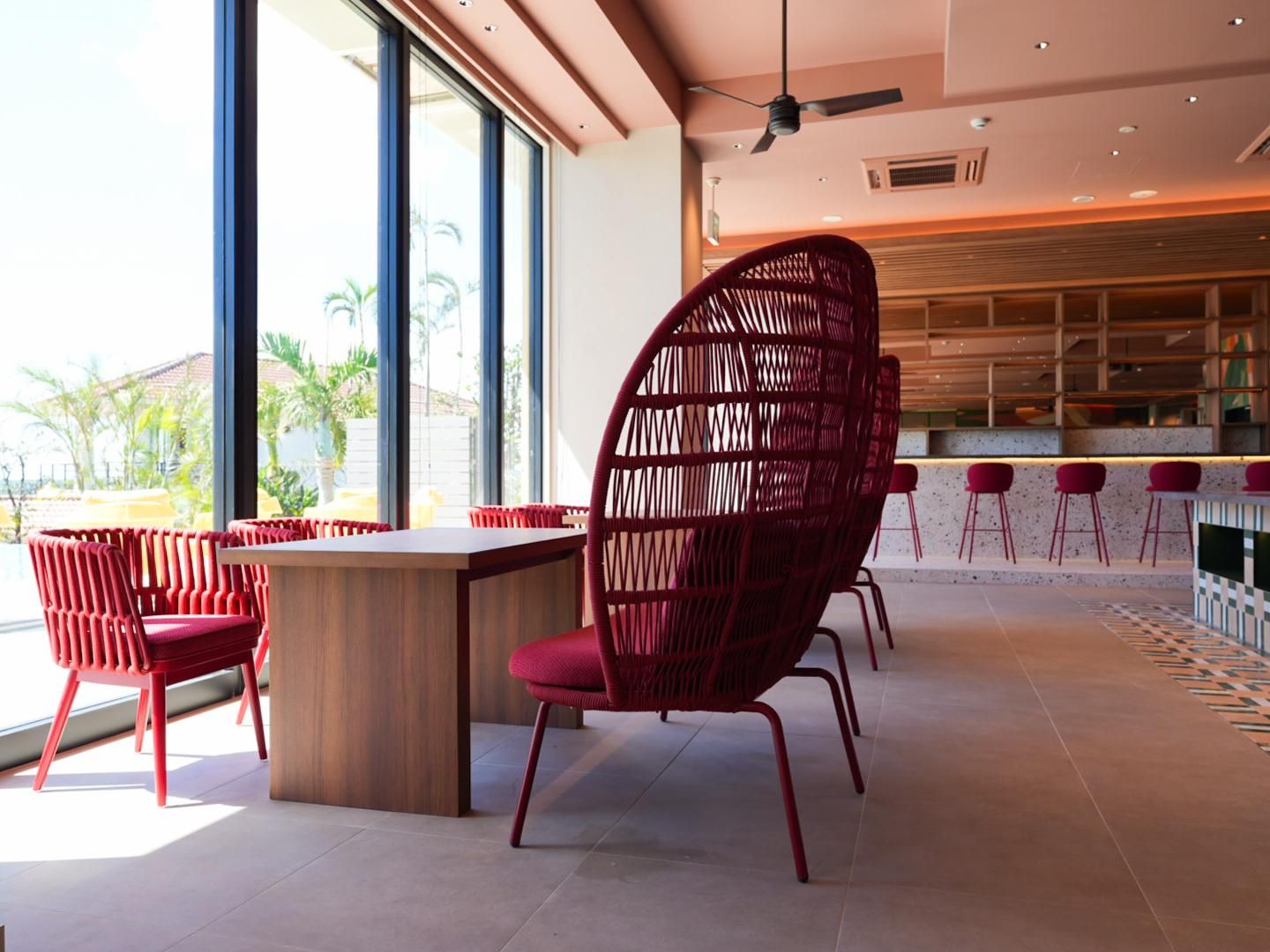 Un interior moderno con comedor que incluye sillas rojas y una mesa de madera, ventanas amplias con vista a palmeras y un área de bar con taburetes rojos.