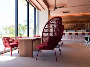 Un interior moderno con comedor que incluye sillas rojas y una mesa de madera, ventanas amplias con vista a palmeras y un área de bar con taburetes rojos.