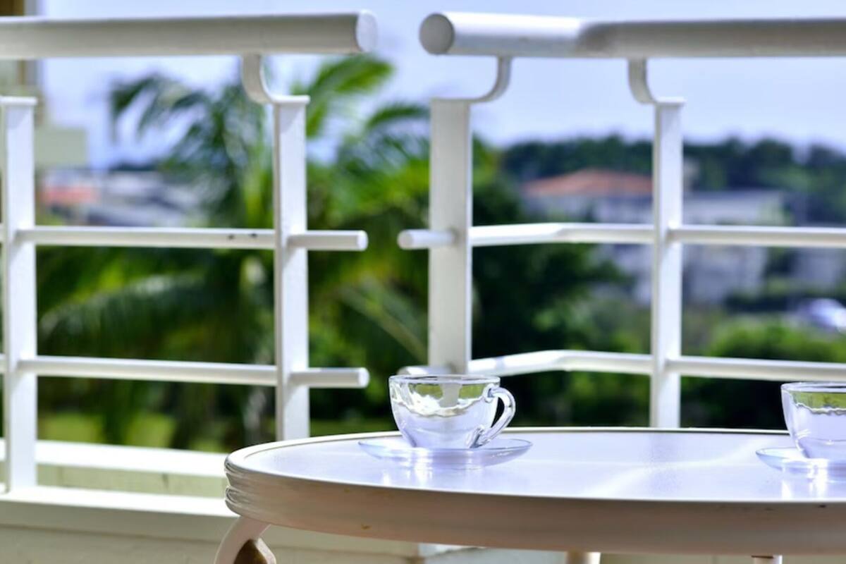 A balcony with a white railing, a round table with two clear glass cups, and a view of greenery and buildings in the background.