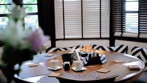 A round dining table with a black and white patterned cushion, two white cups, a black mug, and a basket of bread.