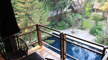 A wooden deck with a pool, surrounded by tropical vegetation and a small house.