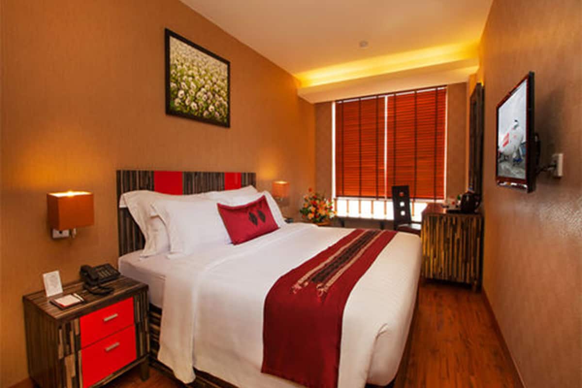 A hotel room with a large bed, a bedside table with a lamp, a small table with a vase of flowers, and a painting on the wall.