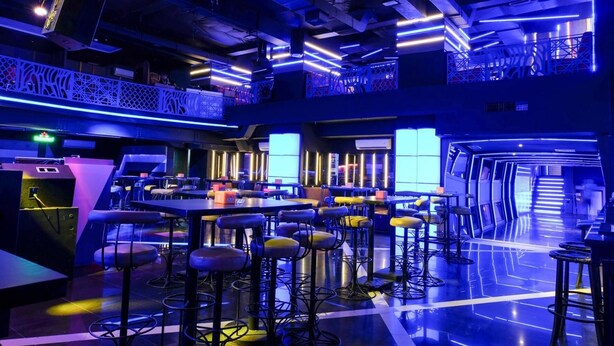A modern bar area with multiple seating options, including high stools and tables, under blue ambient lighting. - Almadera Hotel Makassar (Makassar)