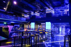 Zona de bar moderna con diversas opciones de asiento, incluyendo taburetes altos y mesas, iluminada con luz azul ambiental.