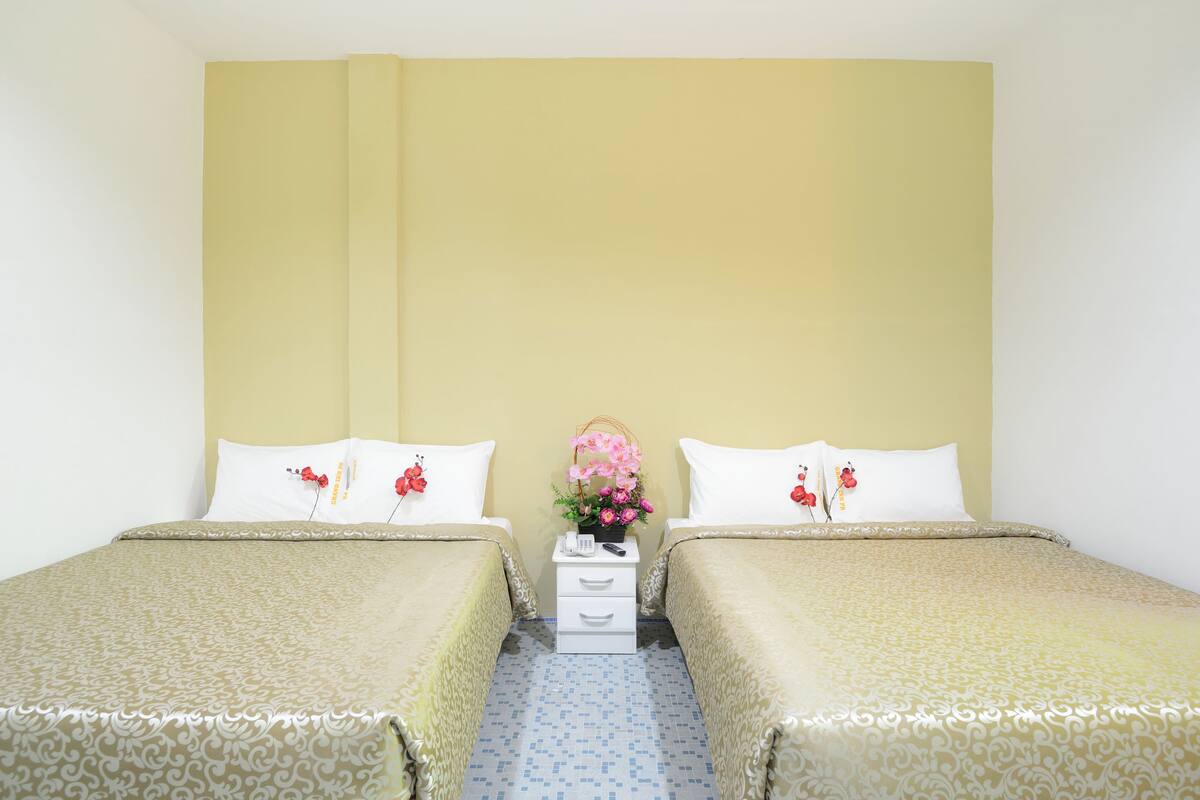 A double room with two beds, a nightstand with a phone and remote, and a small table with a vase of flowers.