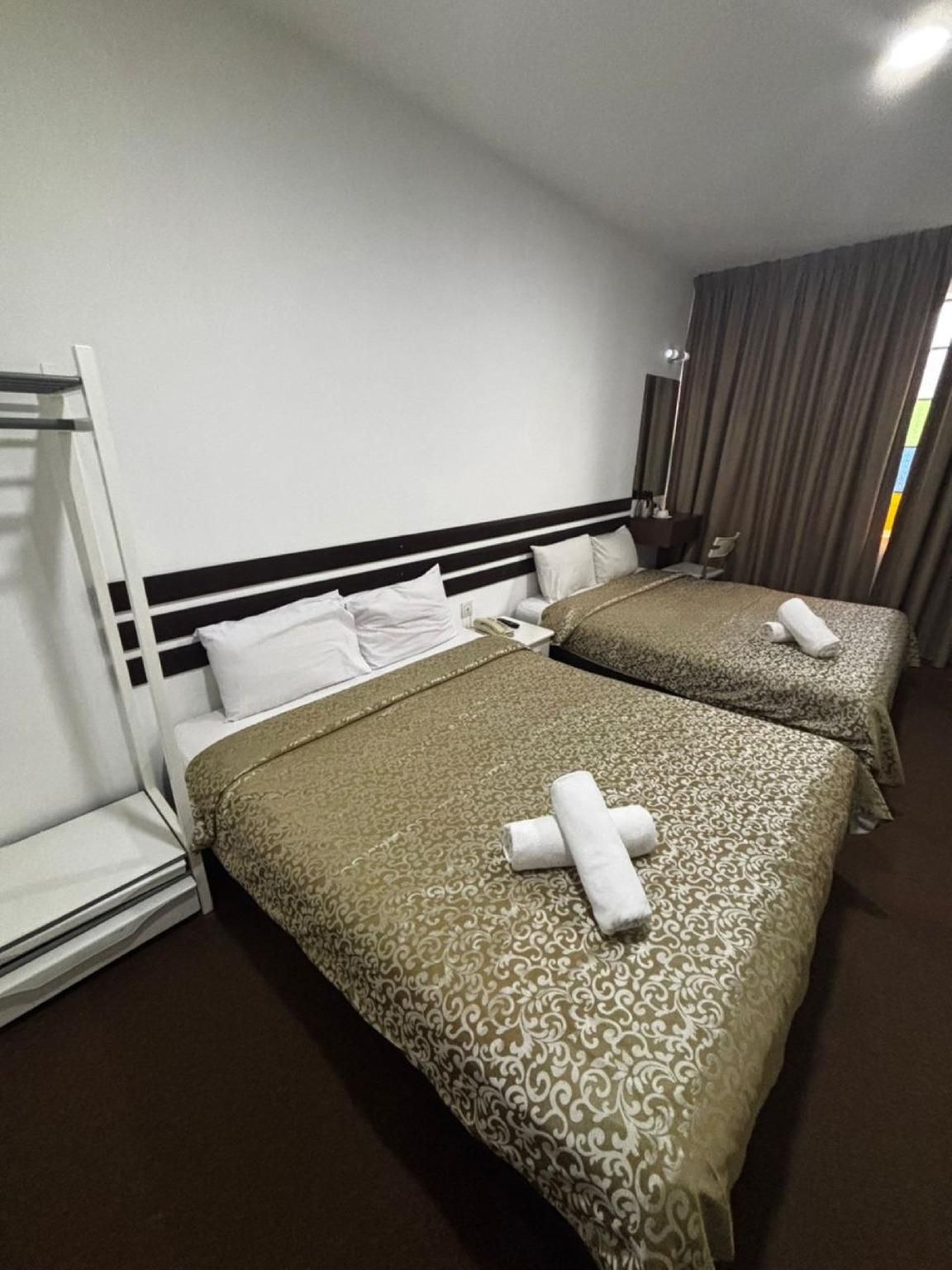 A hotel room with two beds, a headboard, a bedside table, a lamp, and a window with curtains.