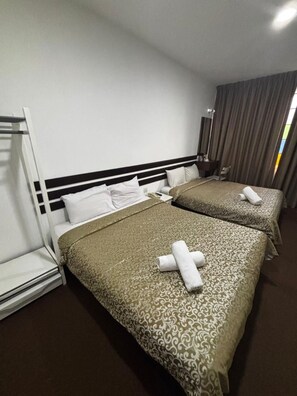 A hotel room with two beds, a headboard, a bedside table, a lamp, and a window with curtains.