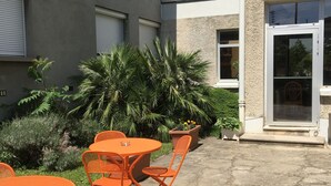 Een terras buiten met oranje metalen tafels en stoelen, omgeven door groen en een gebouw met een stenen gevel.