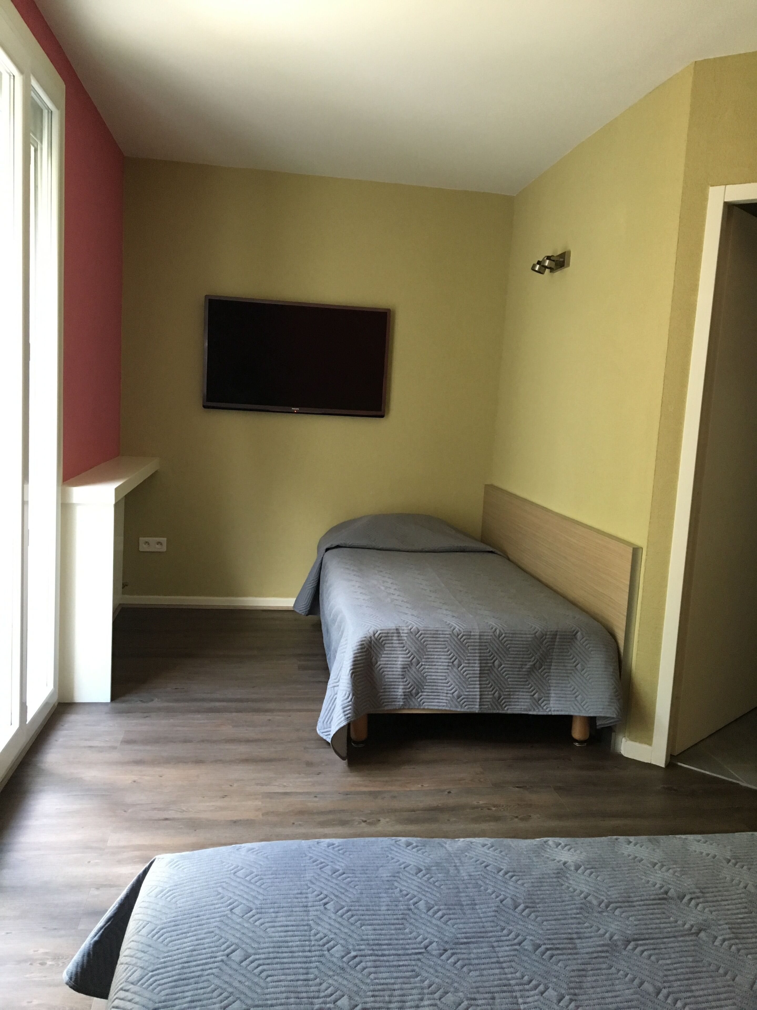 Una piccola camera con un letto singolo, televisione a parete, pavimento in legno e una finestra che fa entrare luce naturale.