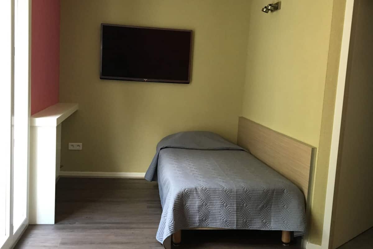 Quarto pequeno com uma cama de solteiro, televisão na parede, piso de madeira e janela que permite a entrada de luz natural.