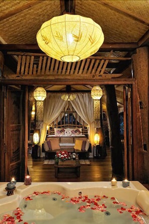A traditional room with a wooden ceiling, a large hanging lamp, a bed with a canopy, a bathtub with rose petals, and candles. - Fern Paradise (San Sai)