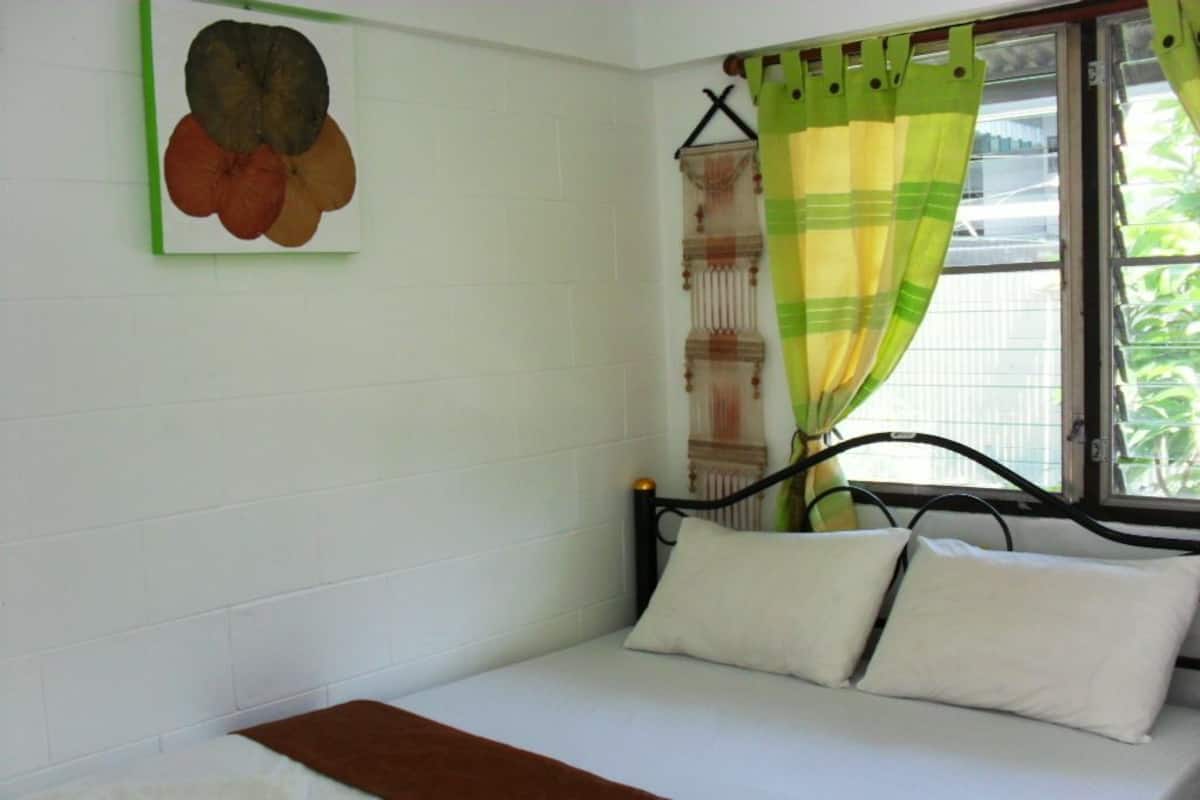 A single bed with white bedding and a brown throw, a green and yellow striped curtain, and a wall-mounted painting of fruits.