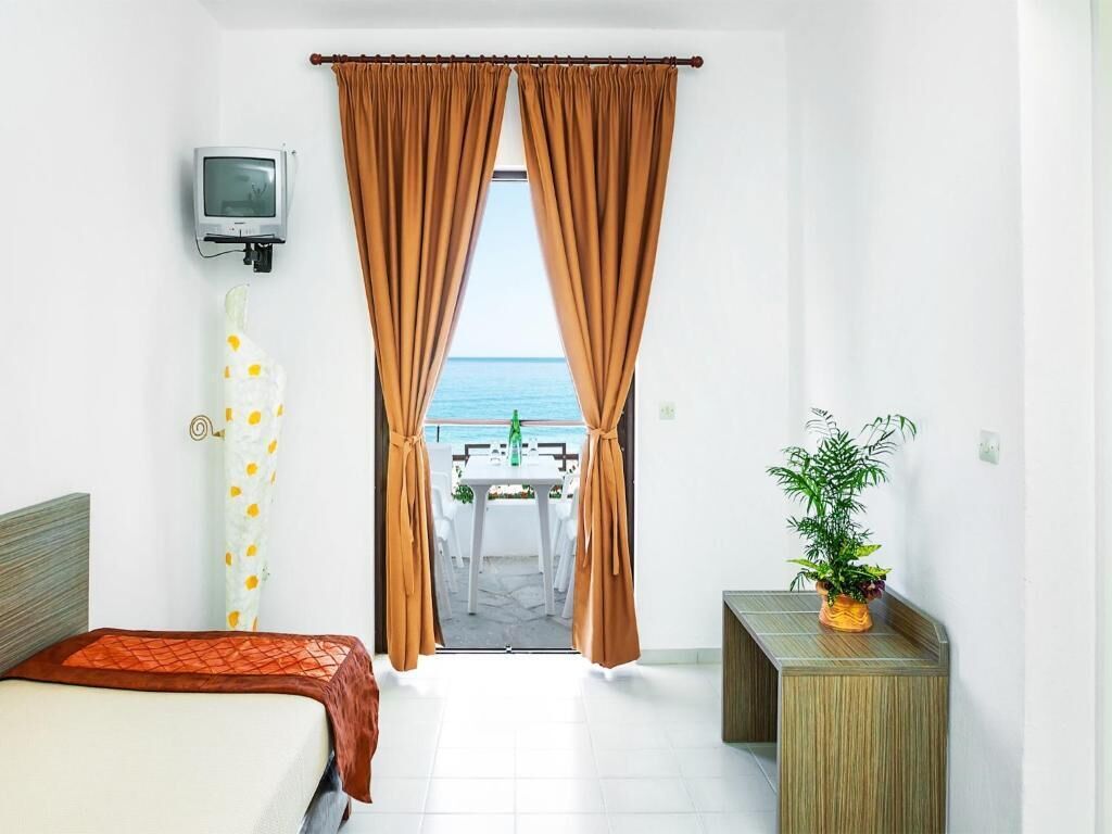 A hotel room with a bed, a small table, a potted plant, and a view of the sea through curtains.