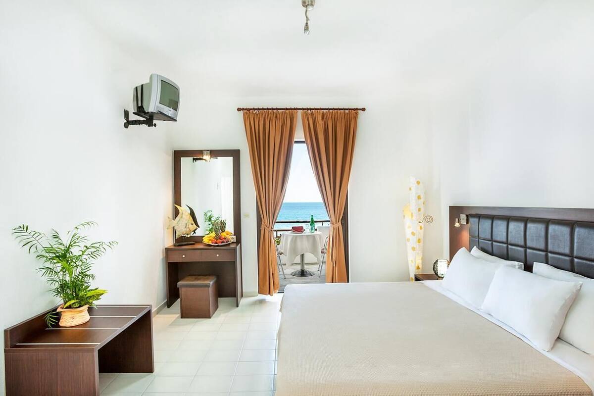 Ein Hotelzimmer mit Bett, Schreibtisch, Stuhl, einer Zimmerpflanze, einem Spiegel und Meerblick durch Vorhänge.