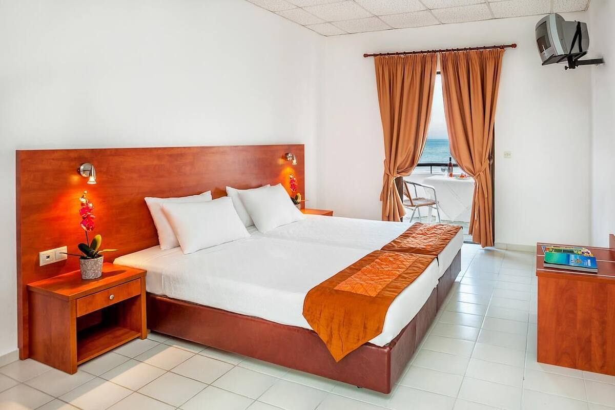 Ein Hotelzimmer mit Holz-Kopfteil, einem Bett mit weißer Bettwäsche, einem Nachttisch mit Lampe und Pflanze, einem Schreibtisch mit Stuhl und Blick auf das Meer durch Vorhänge.