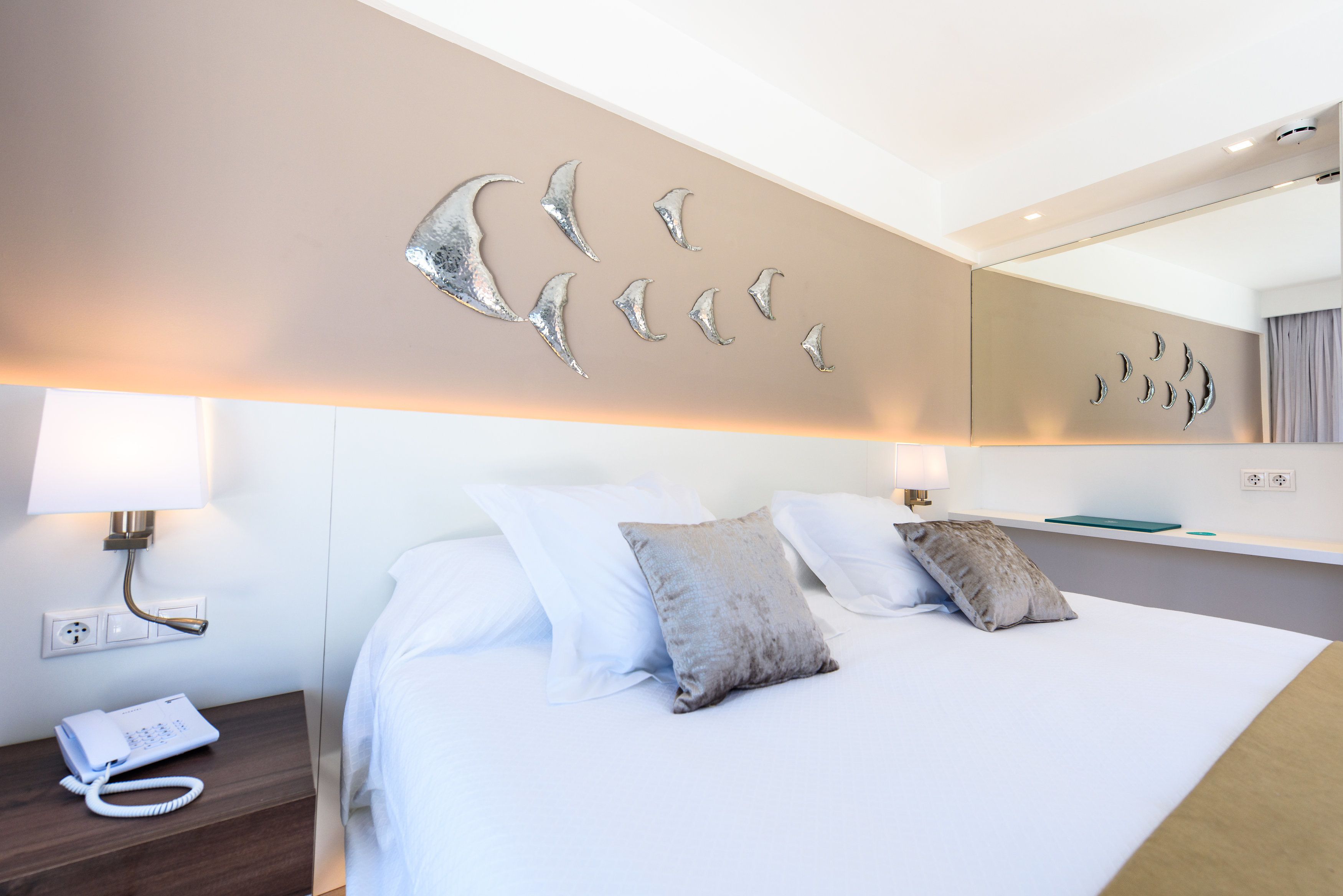 A hotel room with a bed, a nightstand with a phone, a wall-mounted light, and a mirror.