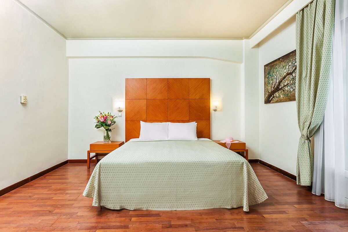 A hotel room with a wooden headboard, a bed with a green bedspread, a nightstand with a vase of flowers, and a painting on the wall.